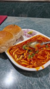Mumbai-style misal pav with spicy red-orange curry, bread rolls, onions, coriander, and lime.
