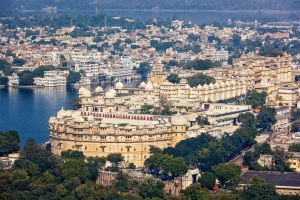 Udaipur Palace by the Lake: A large historic palace complex stands along the waterfront, featuring ornate domes, arches and sandstone-colored façades, with a small red boat on Lake Pichola.