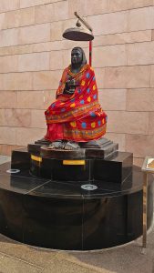 A statue of Ahilya Bai Holkar seated beneath a decorative umbrella, draped in a vibrant red saree with colorful patterns and adorned with flower garlands, displayed on a black stone pedestal against a beige stone wall.
