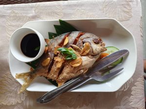 A golden fried whole pompano fish topped with crispy garlic chips and scallions, served with a ramekin of dark soy dipping sauce and cucumber slices on a white platter.