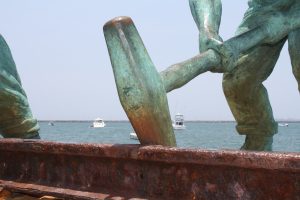 Close-up of a green-patinated bronze sculpture showing a worker holding a large mallet over a rusty iron rail, with boats on the sea in the background under a clear sky.