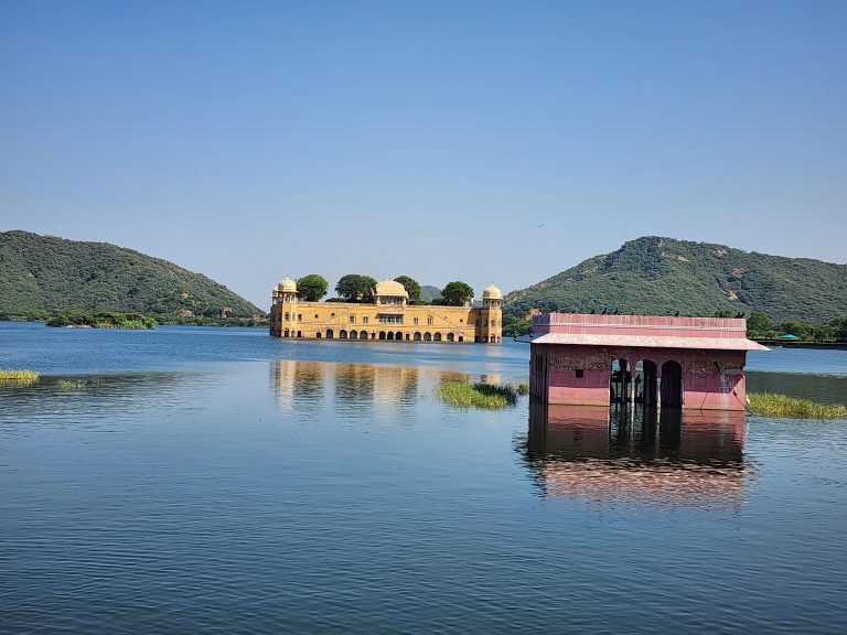 Jal Mahal is in the middle of Man Sagar Lake in the city of Jaipur, Rajasthan, A serene landscape featuring a large, yellow architectural structure rising from a calm body of water, surrounded by rolling green hills. The building has arched openings and is topped with distinctive domes and lush trees. In the foreground, a smaller, pink pavilion partially submerged in water reflects in the lake, with delicate details visible on its walls. The clear blue sky and gentle ripples add to the tranquil scene, creating a harmonious connection between nature and human-made beauty.