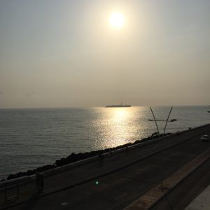Panoramic view of a coastal boardwalk at sunset, with sunlight reflecting on the ocean and the silhouette of an island or vessel on the distant horizon.