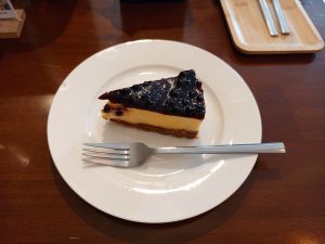 A slice of baked blueberry cheesecake with a thick fruit compote topping and a golden graham crust, served on a white dessert plate with a fork.