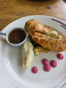 A plated gourmet chicken dish, appearing to be a stuffed chicken ballotine or roulade served with mashed potatoes, roasted vegetables, a small pot of jus or gravy, and pink puree dots on the side