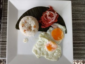 A traditional Filipino cuisine cornsilog breakfast plate featuring sautéed corned beef topped with sliced red onions, a scoop of garlic rice, and two sunny-side-up fried eggs on a banana leaf.

