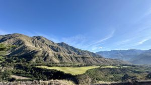 Wide-angle view of rugged, textured mountains with sparse vegetation and a patch of cultivated green field, under a large blue sky.