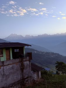 A concrete and brick building in the foreground overlooking a distant valley and a snow-capped mountain range under a pale morning sky.