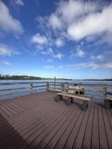 A wooden dock extends over a calm body of water, with a railing enclosing the area. The dock is made of dark-red planks, and there is a simple wooden bench on the left side. In the background, the water reflects the blue of the sky, which is dotted with white, fluffy clouds. Along the edge of the dock, there is a sign indicating caution.