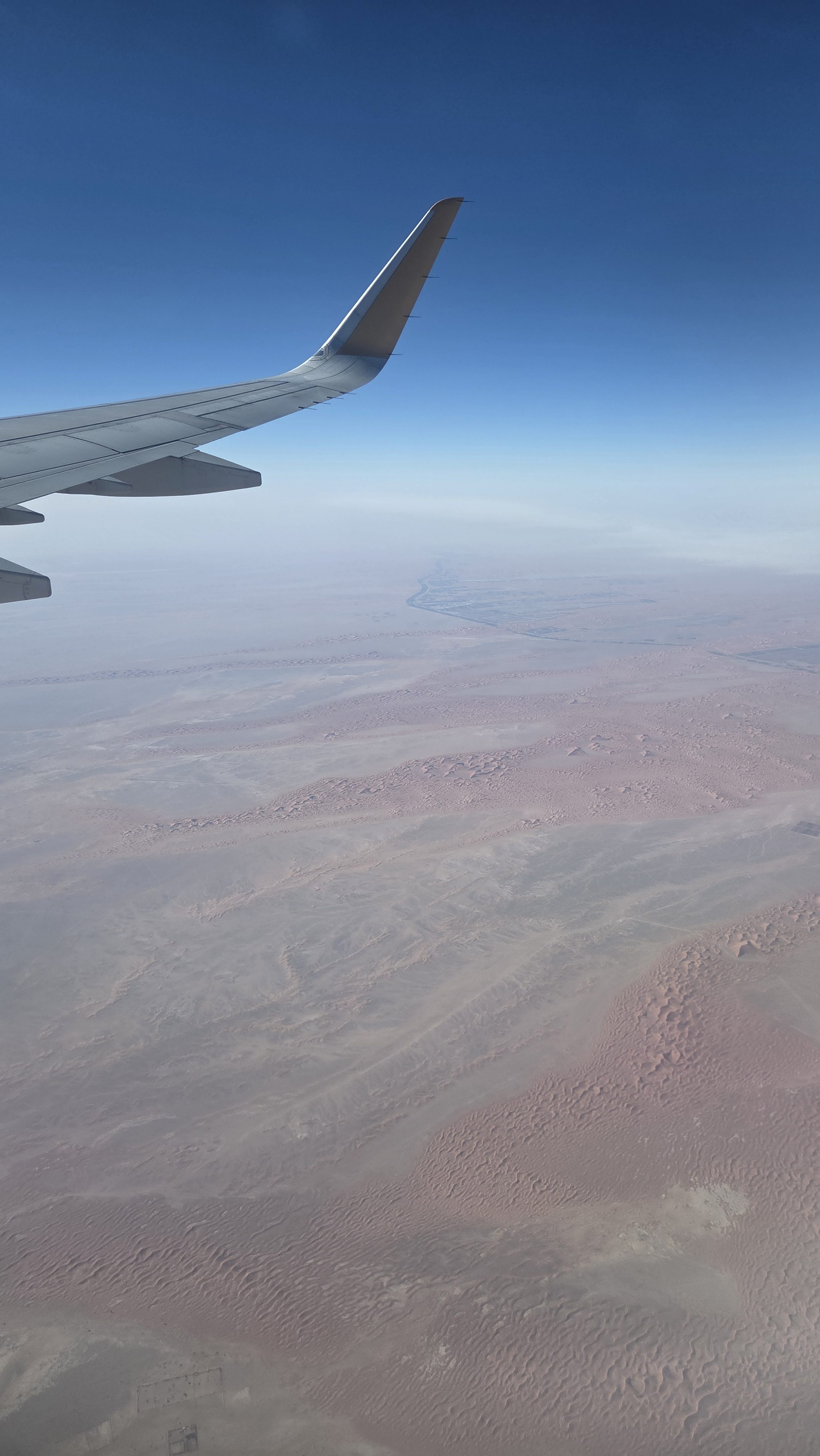 An aerial view from an airplane window looking down at a vast, arid desert landscape under a clear blue sky.