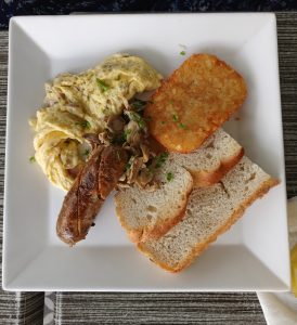 A breakfast plate containing folded scrambled eggs with herbs, sautéed sliced mushrooms, a browned breakfast sausage, a crispy golden hash brown, and thick slices of crusty bread.