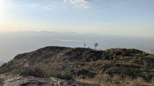A rocky hillside with sparse vegetation overlooks a hazy valley and distant mountain ranges under a clear sky.