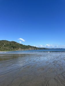 Sandy beach with blue ocean water, white clouds, green hills, and a crescent moon in the sky.