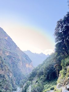 A high-angle view looking down a lush green mountain valley with a winding dirt trail along a river, framed by dense trees and distant mountain silhouettes.