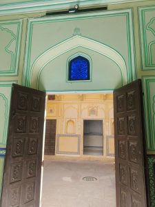 Ornate Rajasthani doorway with carved wooden doors, painted arches, decorative panels, and a blue lattice window.