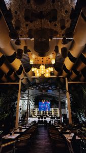 A spacious restaurant interior with a high ceiling adorned with decorative elements. The ceiling features wavy fabric panels in shades of beige and dark colors, creating a dynamic visual effect. Hanging from the ceiling are unique woven pendant lights that emit a warm glow, complemented by circular lights in a cluster. Numerous tables with simple modern design are arranged neatly across the floor, each set with water bottles and condiments. At the far end of the space, a large screen displays a vivid image, and a well-stocked bar is lined with shelves showcasing various bottles. Lush green plants are positioned throughout, adding a touch of nature to the ambiance. The overall atmosphere appears inviting and lively, suggesting a social gathering place.
