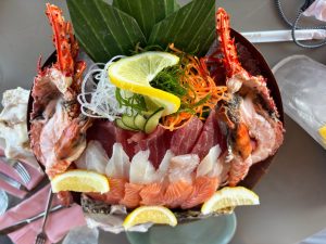 A colorful sashimi platter with salmon, tuna and white fish arranged in rows, flanked by two split lobster halves. Shredded daikon, seaweed, carrot, cucumber, lemon slices and large green leaves decorate the seafood display.