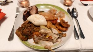 A white ceramic dinner plate filled with assorted dishes including steamed buns, dumplings, fried fish, curry meat, and onion rings set on a formal dining table.
