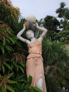 A classical style garden statue of a woman draped in an apricot-toned robe holding a round globe lamp above her shoulder.